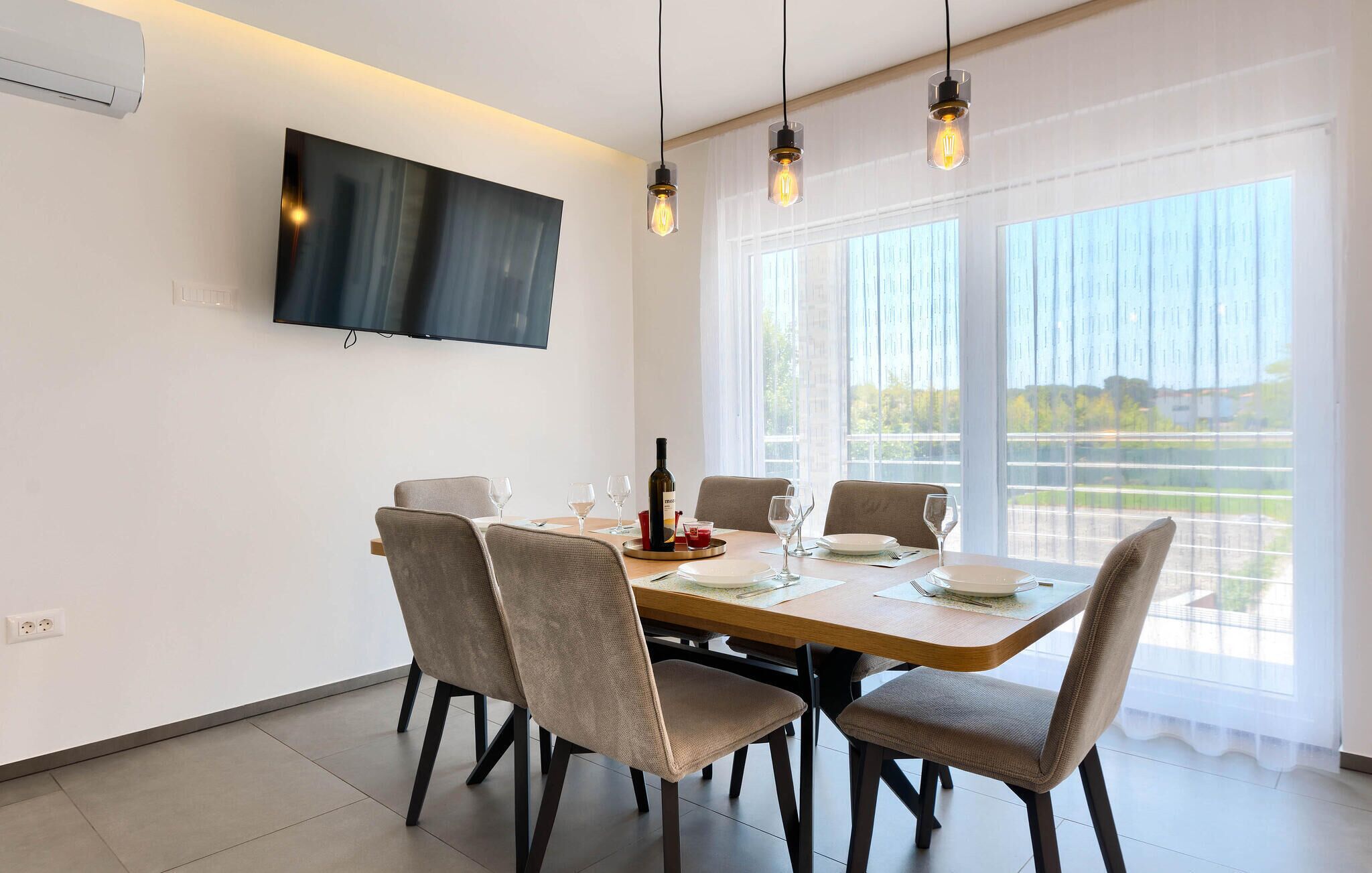 A dining area with a large table, six chairs, a flat-screen TV mounted on the wall, and a view of a green landscape through the windows.