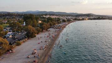 A coastal beach with people sunbathing and swimming, a parking lot, and a view of the city and mountains in the background.