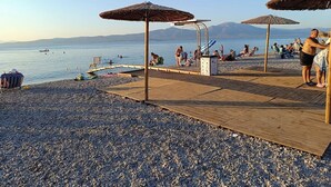 Una spiaggia con ciottoli, passerelle in legno, ombrelloni e zona piscina.