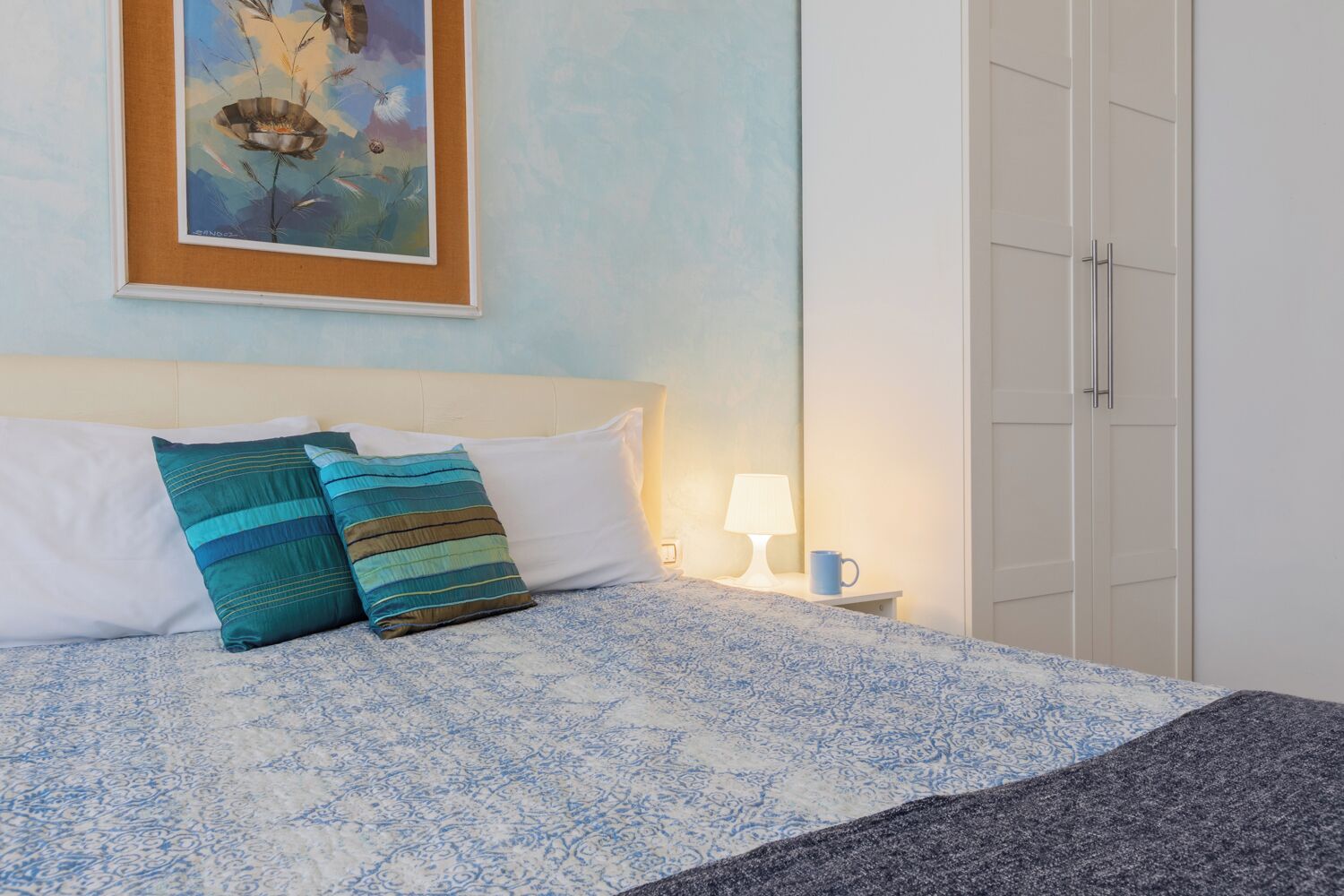 A neatly made bed with a patterned bedspread, two pillows, a bedside lamp, and a framed artwork on the wall.