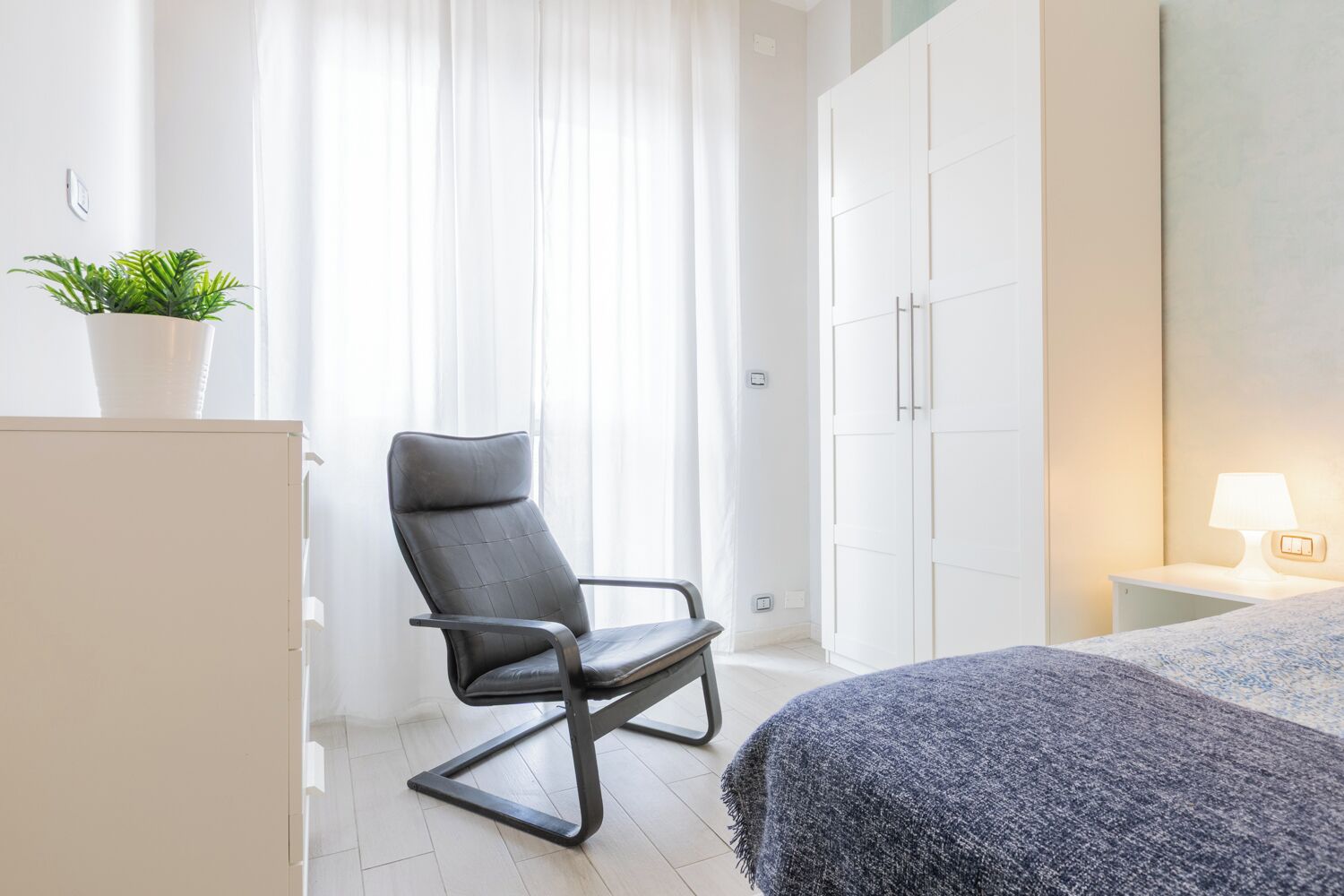 Una habitación de hotel moderna con cama, una silla, un armario y una mesita de noche con lámpara.