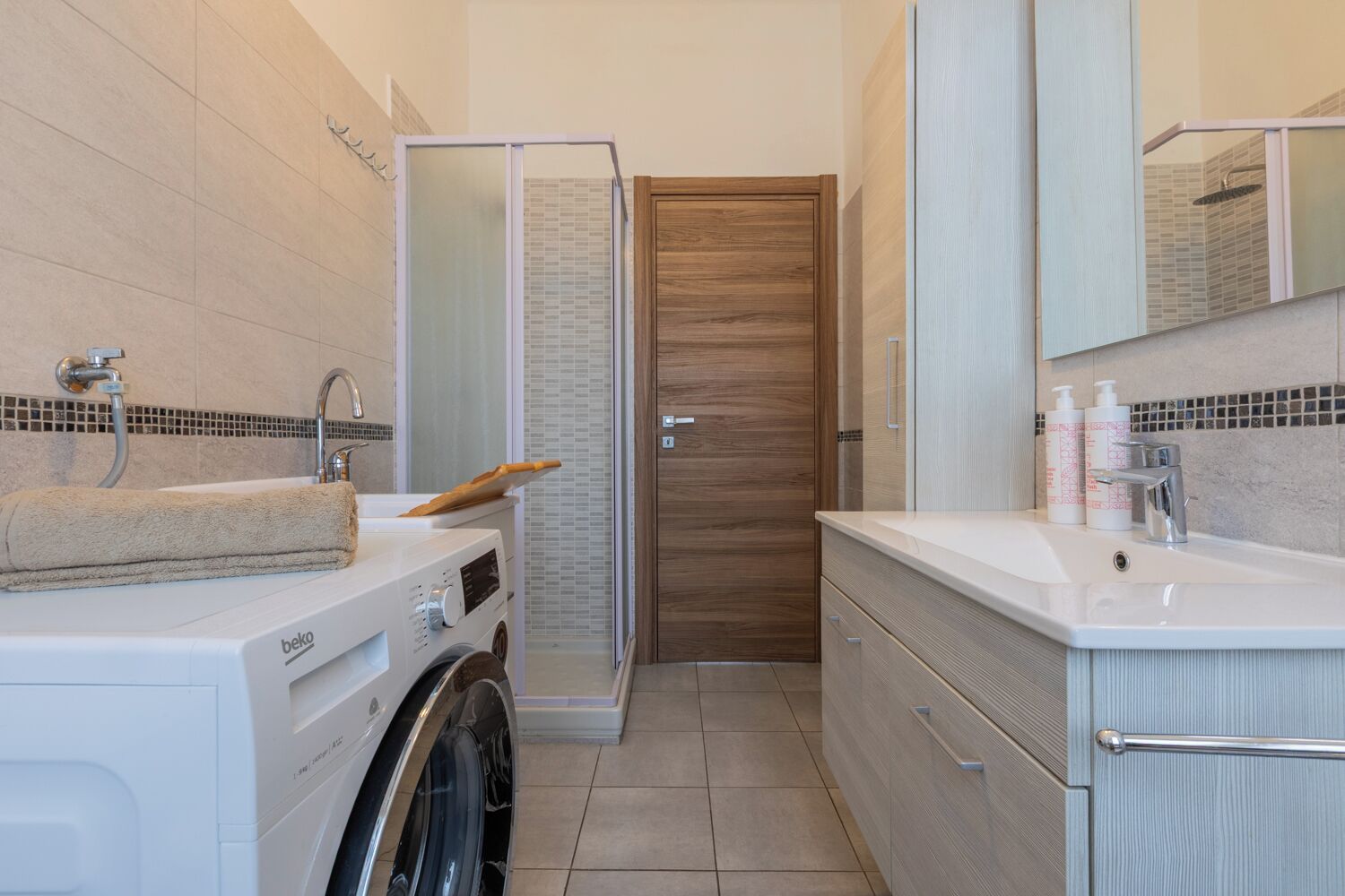 A compact bathroom with a washing machine, a shower enclosure, and a sink with toiletries.