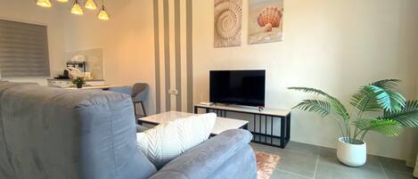 A modern living room with a grey sofa, a flat-screen TV, a dining area with a table and chairs, and a potted plant.