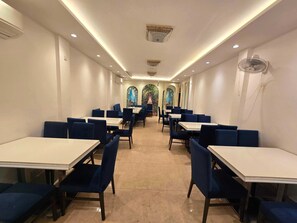 A restaurant with tables and chairs arranged in rows, a ceiling fan, and a view of a garden through arched windows.