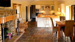 A spacious kitchen with a stone floor, a dining area with a wooden table and chairs, and a stone fireplace. - Howle Hideaway (Ross-on-Wye)