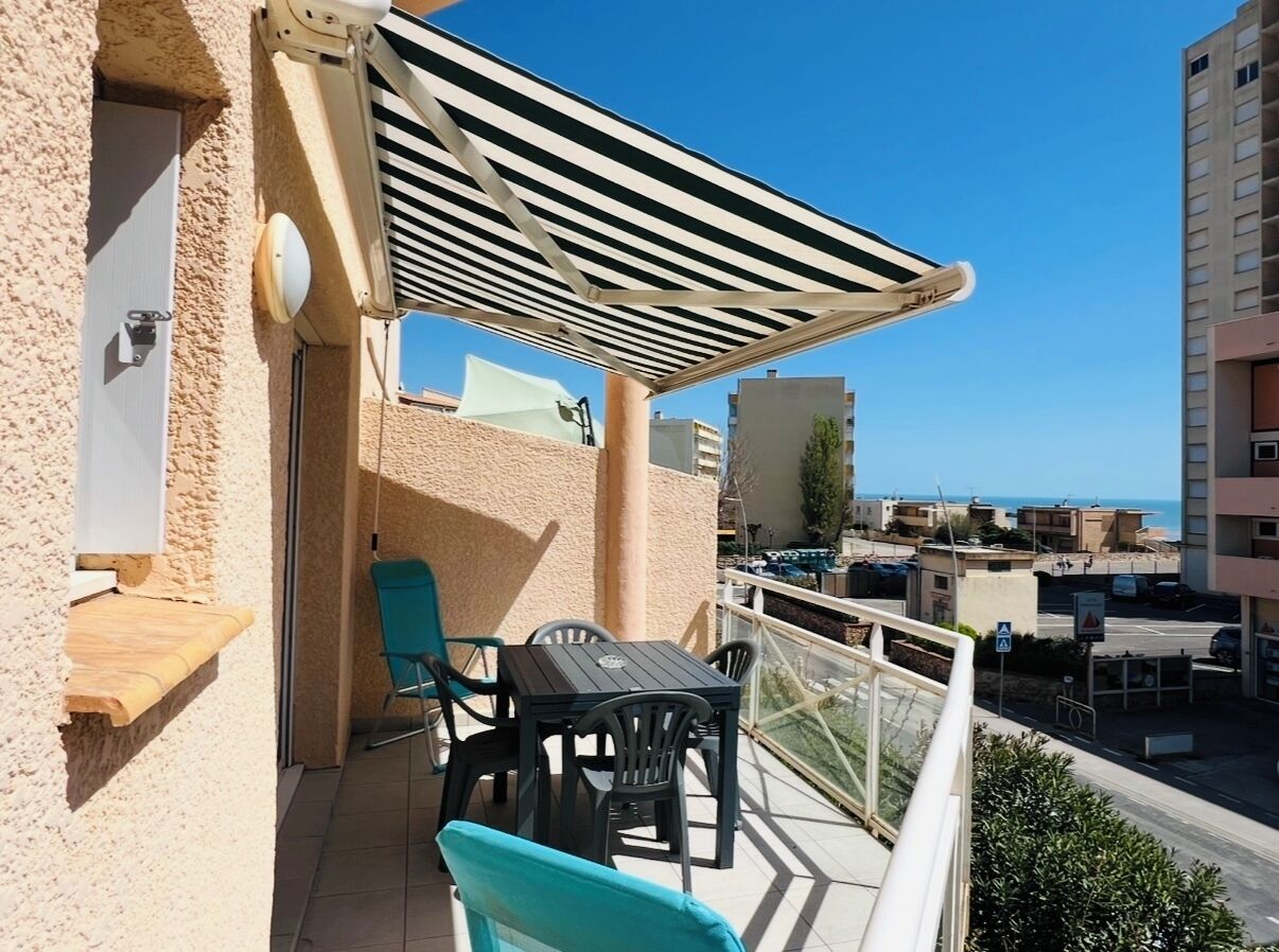 Een balkon met een tafel en stoelen, een houten plank, een gestreepte luifel en uitzicht op zee en gebouwen.