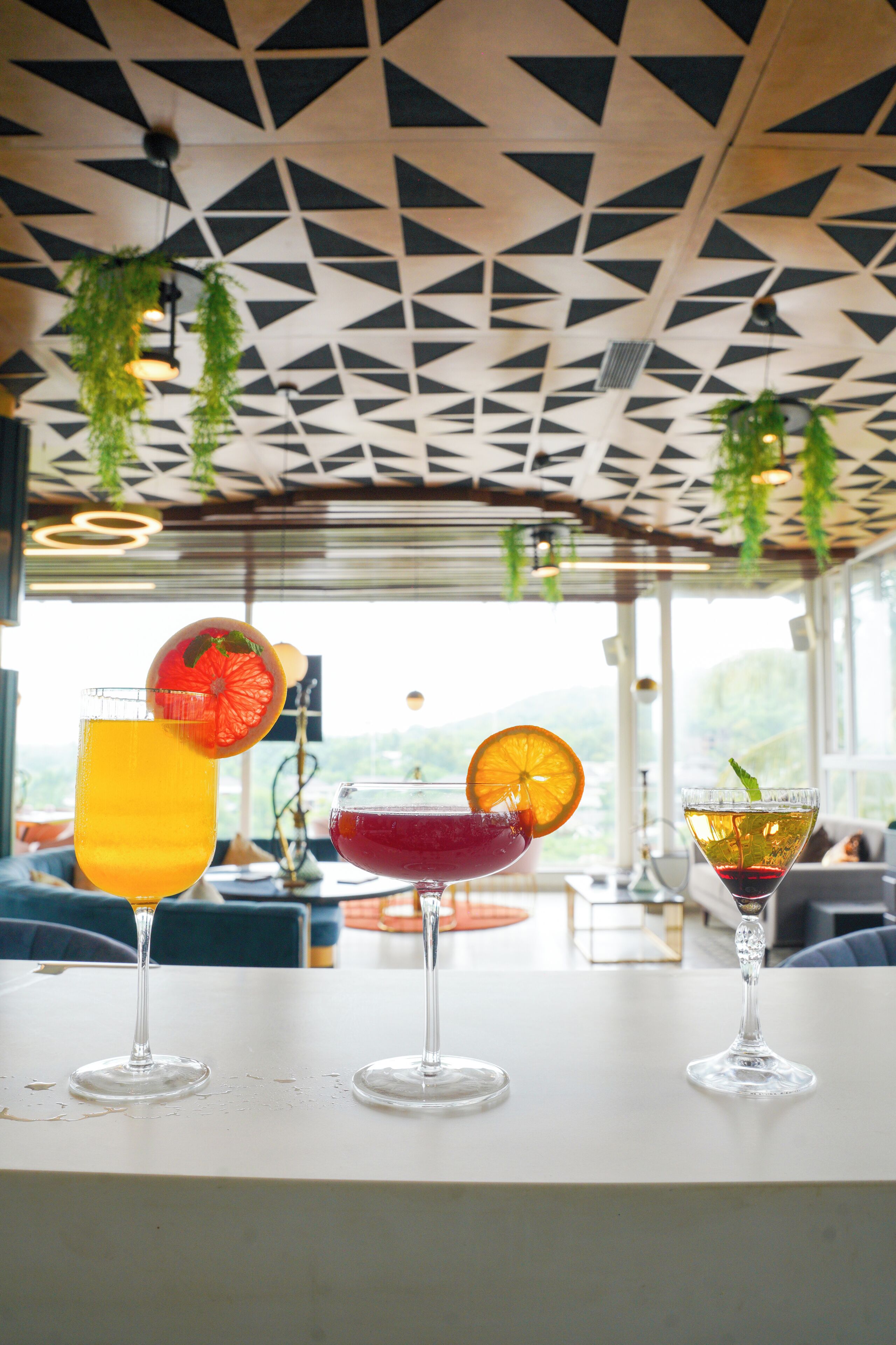 Trois verres de boissons sont posés sur une table, sous un plafond orné de motifs géométriques et agrémenté de plantes suspendues.