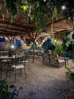 An outdoor seating area with tables and chairs, surrounded by greenery and under a wooden pergola.