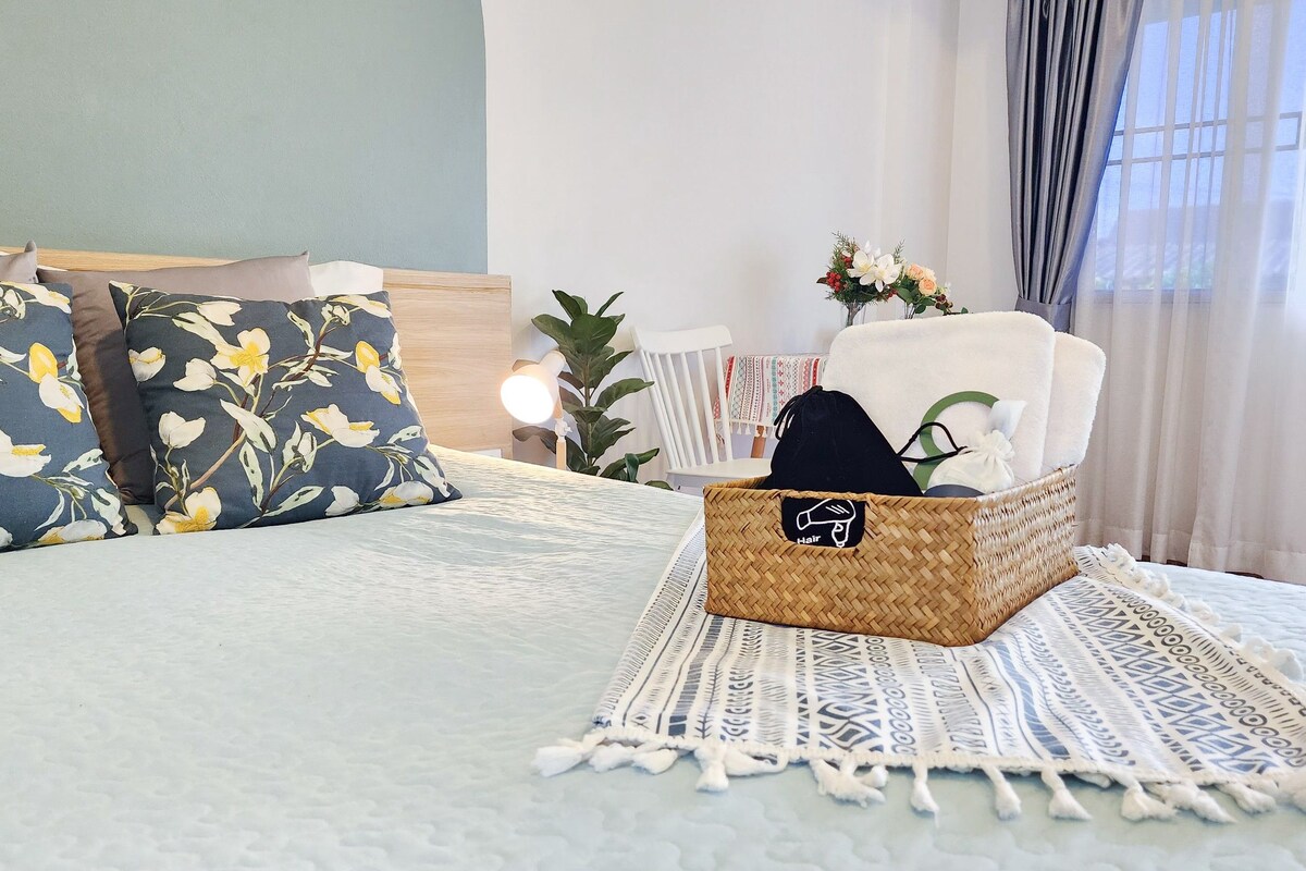 Una cama bien hecha con almohadas estampadas, una cesta tejida con el icono de un cabello y una planta en la mesita de noche.