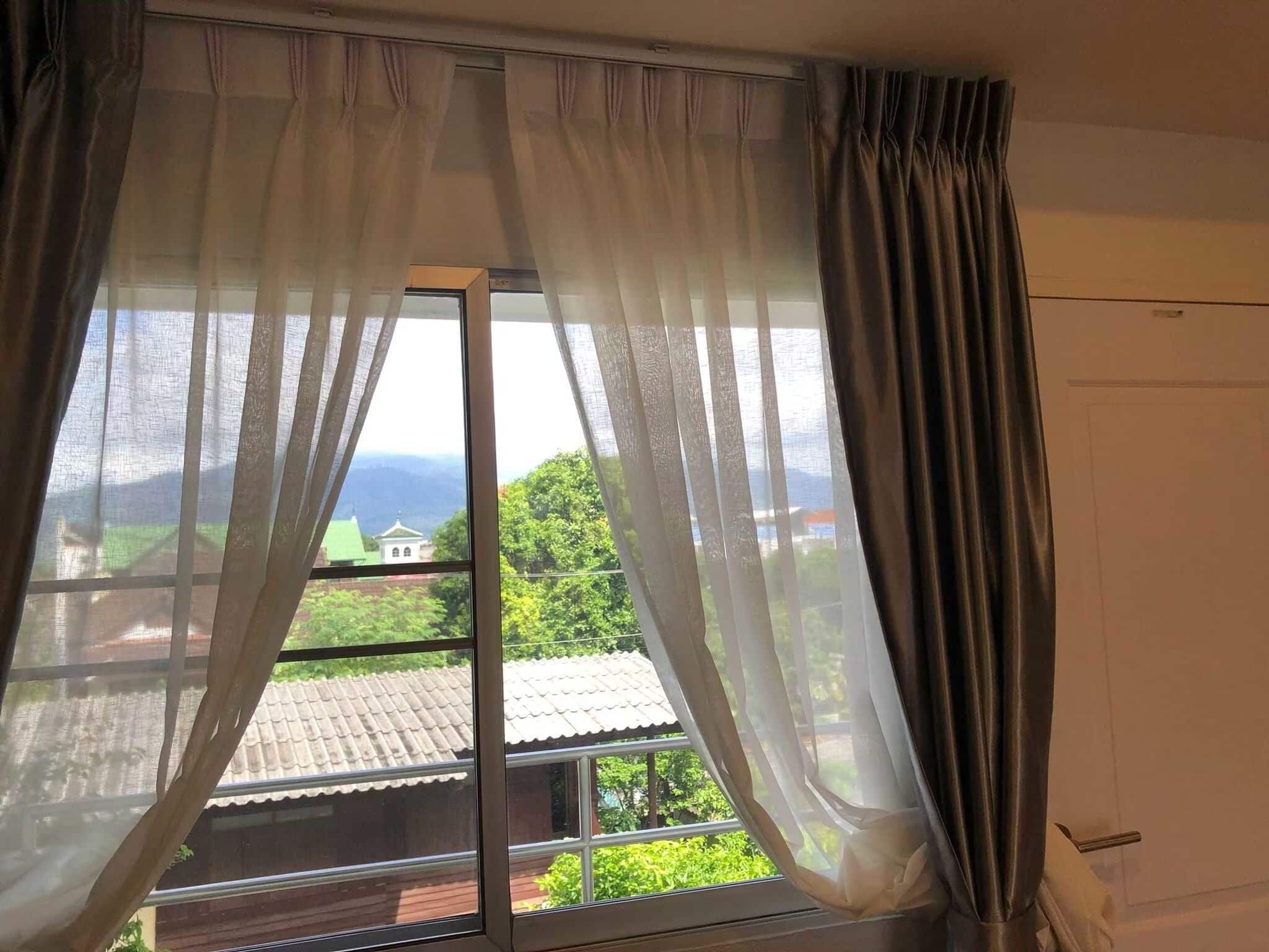 A room with a large window, curtains, and a view of a green landscape with buildings.