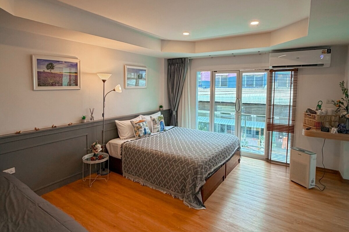 Une chambre moderne avec un grand lit, une table de chevet, une lampe sur pied et une vue sur la ville par la fenêtre.