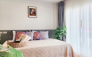 Ein ordentlich bezogenes Bett mit gemusterten Betttextilien, ein Wandbild und ein Fenster mit durchsichtigen Vorhängen.