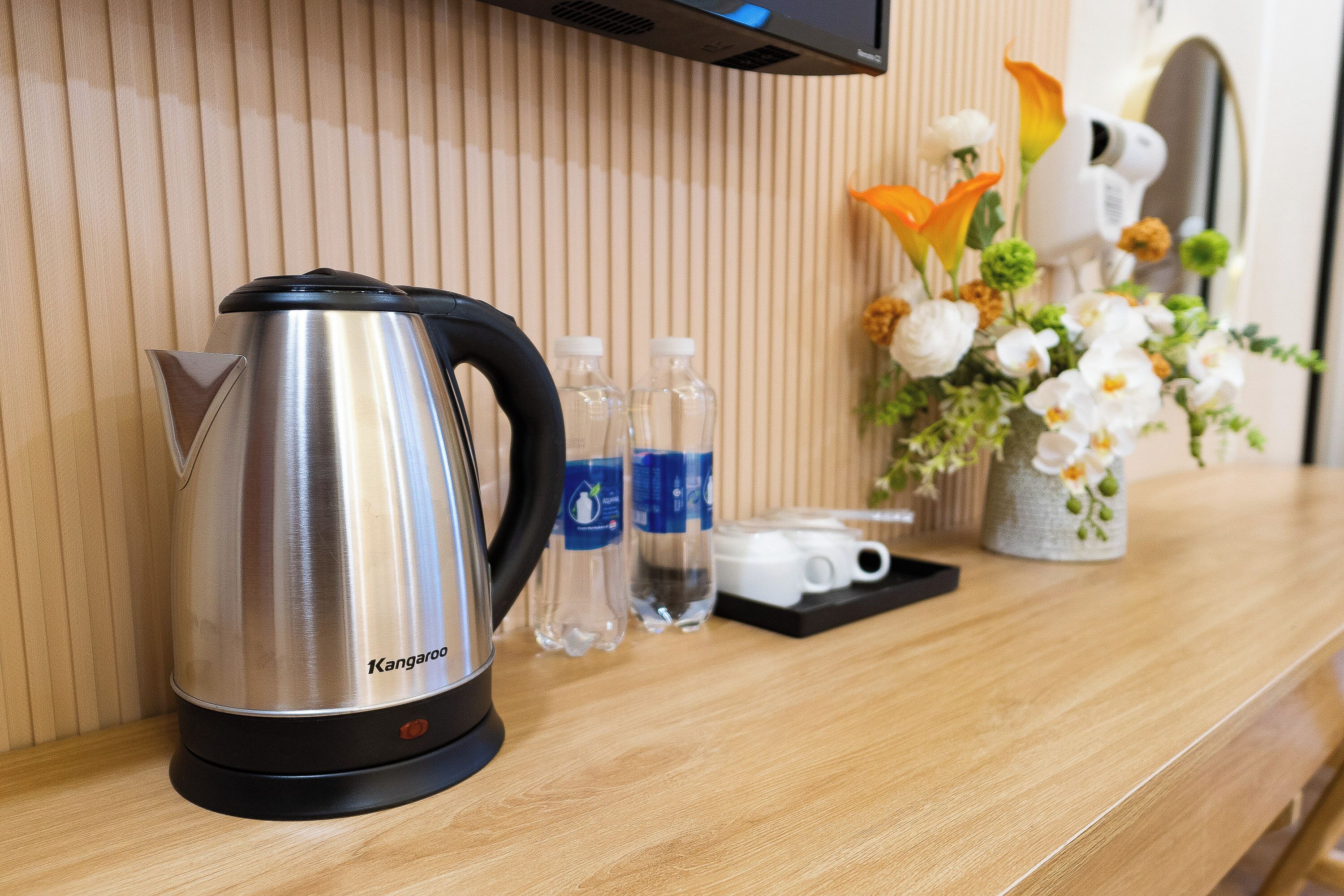A stainless steel kettle on a wooden countertop, clear water bottles, a white mug, and a floral arrangement.