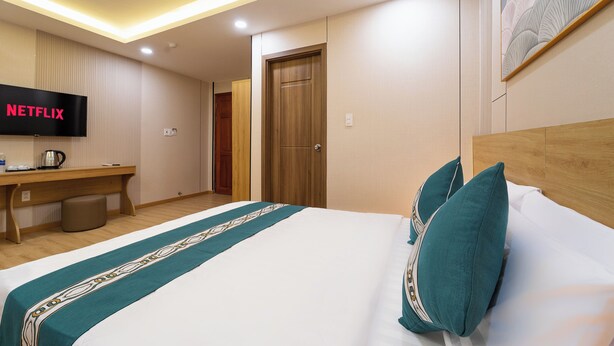 A hotel room with a bed, a desk with a television displaying 'NETFLIX,' a wooden door, and a wall-mounted artwork. - Green Airport Hotel 2 (Ho Chi Minh City)