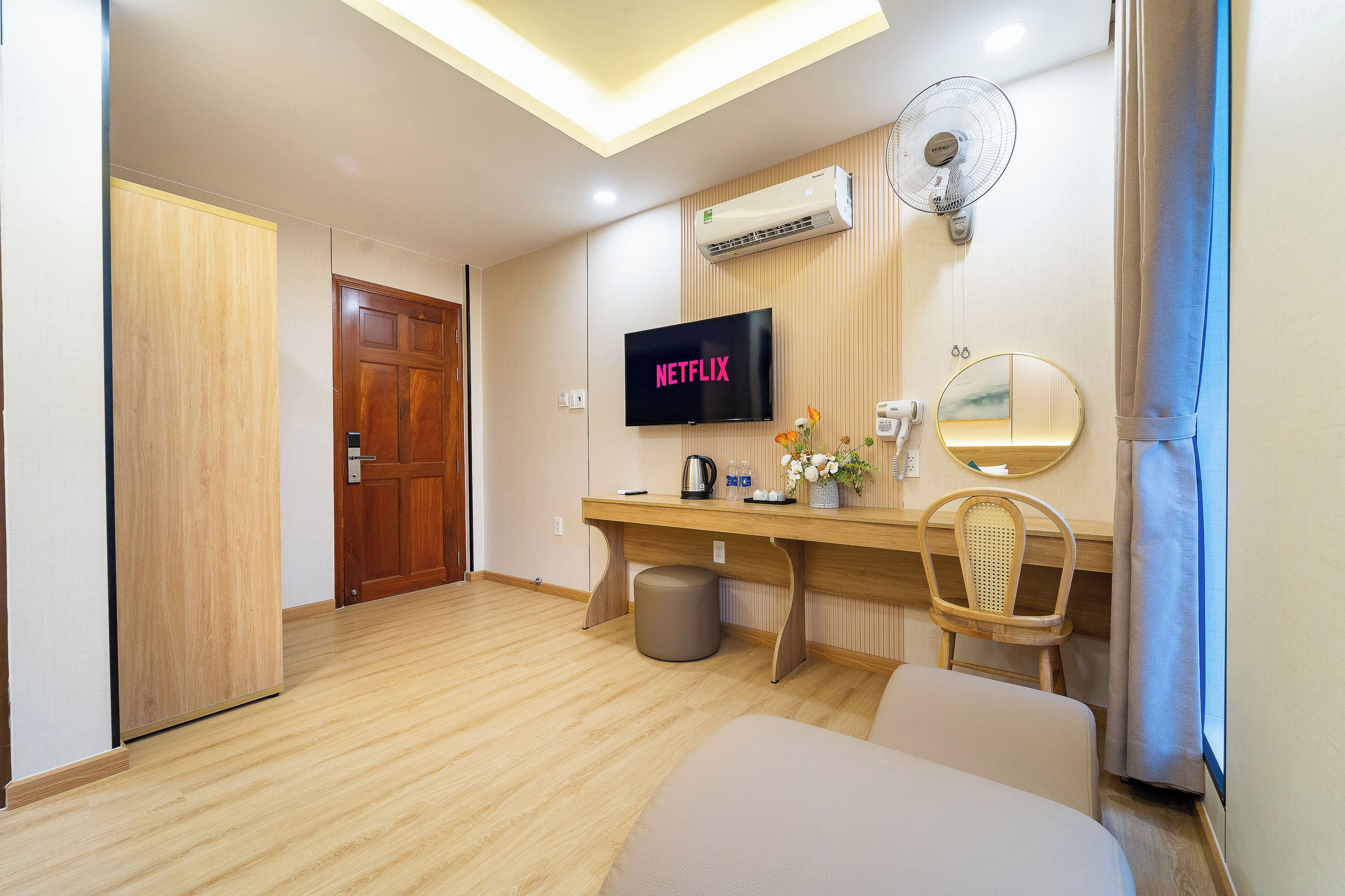 Una camera d'albergo con pavimento in legno, una scrivania con sedia, un televisore che mostra 'NETFLIX', un condizionatore d'aria a muro e una finestra con tende.