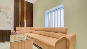 Ein Zimmer mit einem beigefarbenen Sofa, einem Glastisch, einer Wandpaneele aus Holz und einem Fenster mit vertikalen Jalousien.