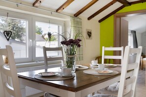 A dining area with a wooden table, white chairs, a vase with flowers, and a window with a heart decoration. - Apartment 'Ferienwohnung Hohes Venn' with Wi-Fi (Monschau)