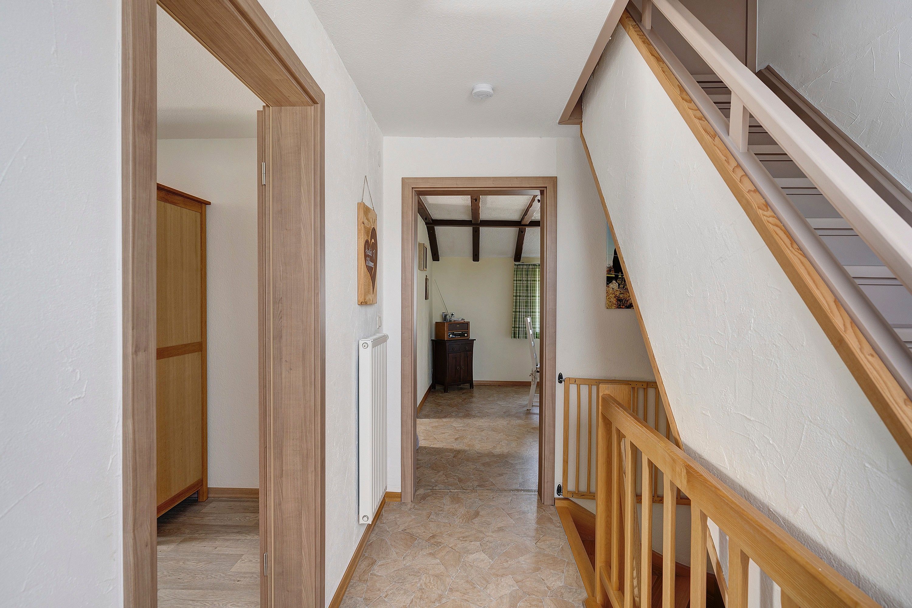 Un pasillo con una escalera de madera, un radiador y una puerta que da a otra habitación.