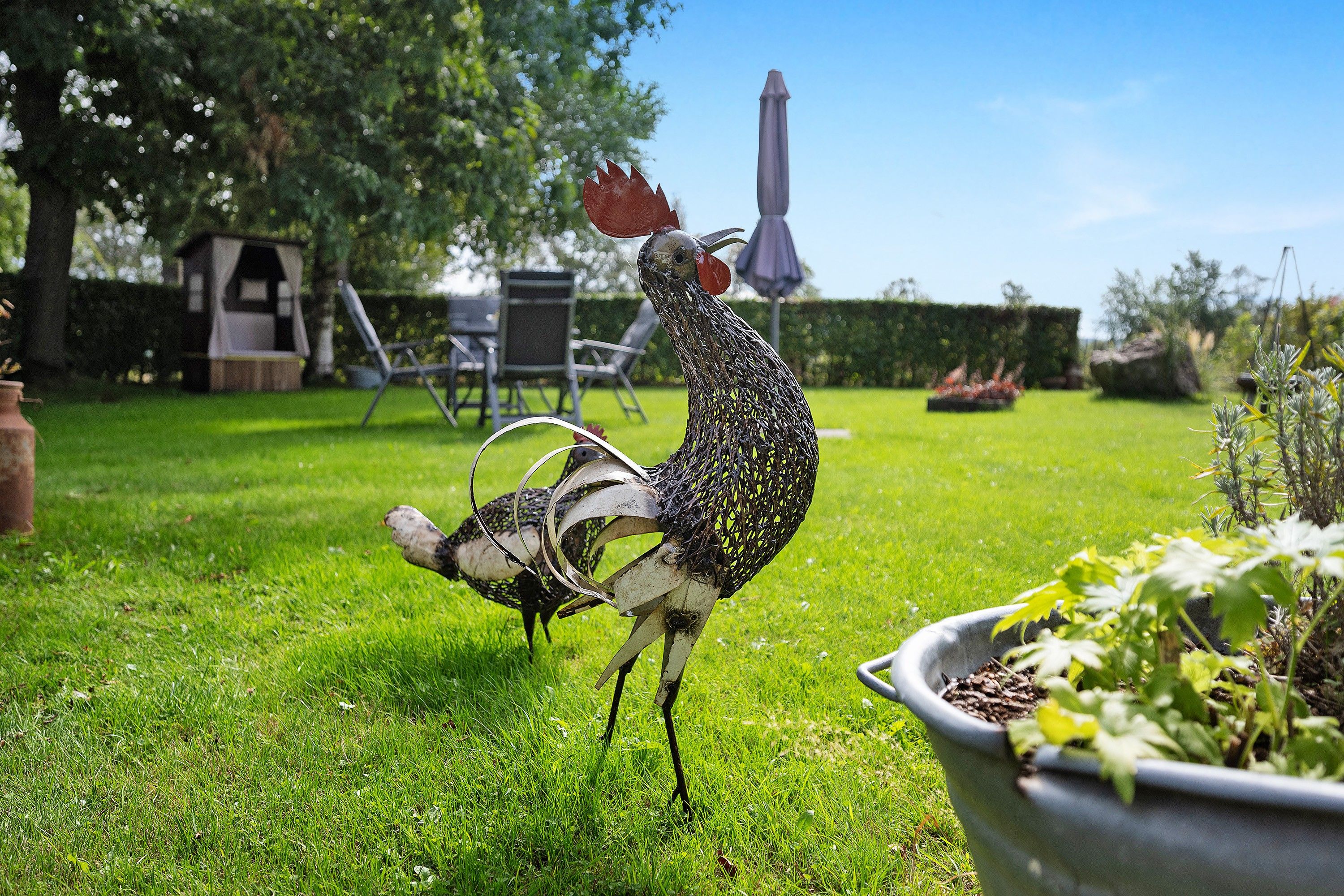 Ein Garten mit einer Vogel-Skulptur aus Metall, einer Rasenfläche und einer Terrasse mit Gartenmöbeln.