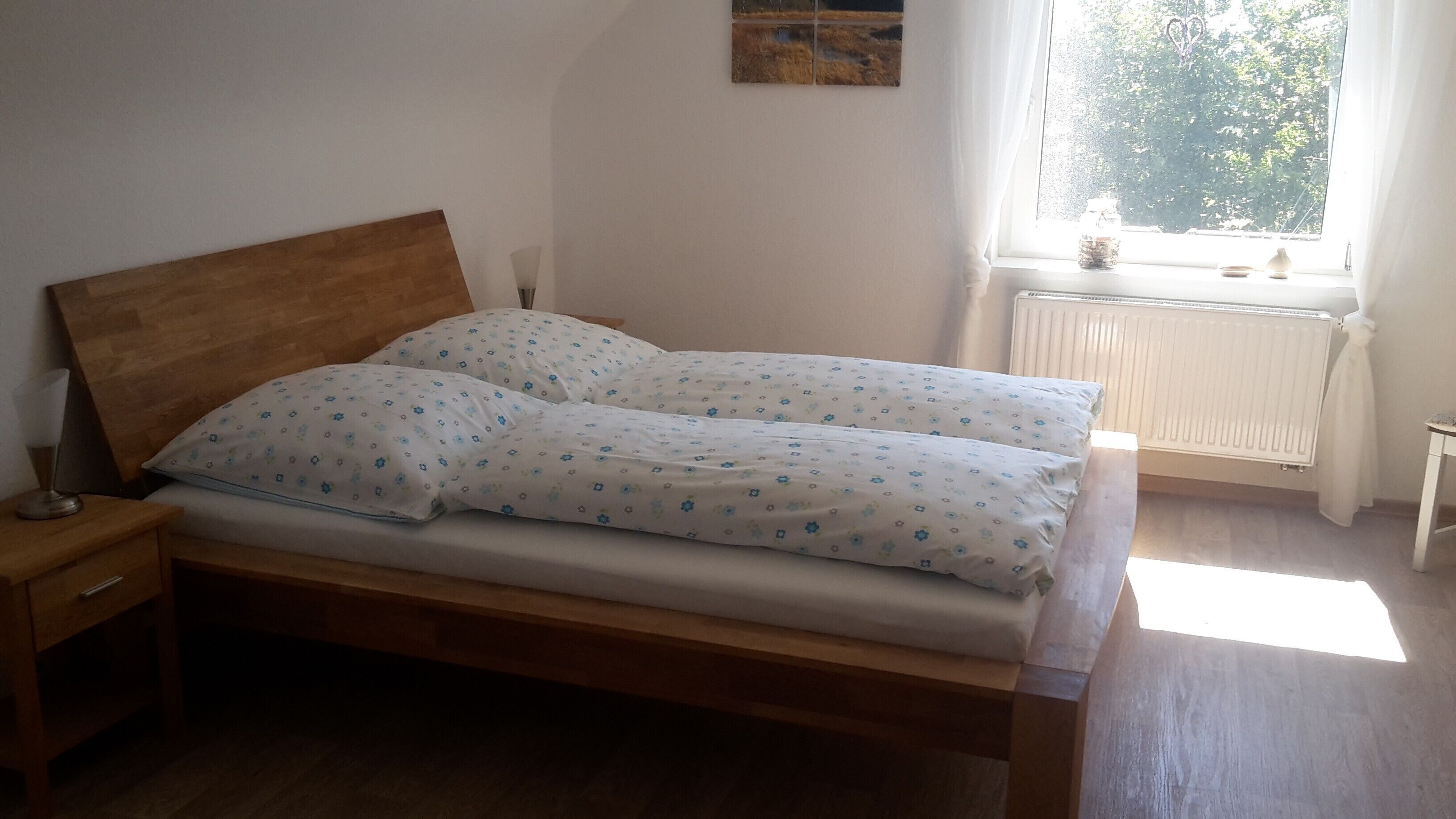 Ein Schlafzimmer mit einem hölzernen Bett, weißer Bettwäsche mit Blumenmuster, einem Nachttisch mit Lampe und einem Fenster mit Blick ins Grüne.