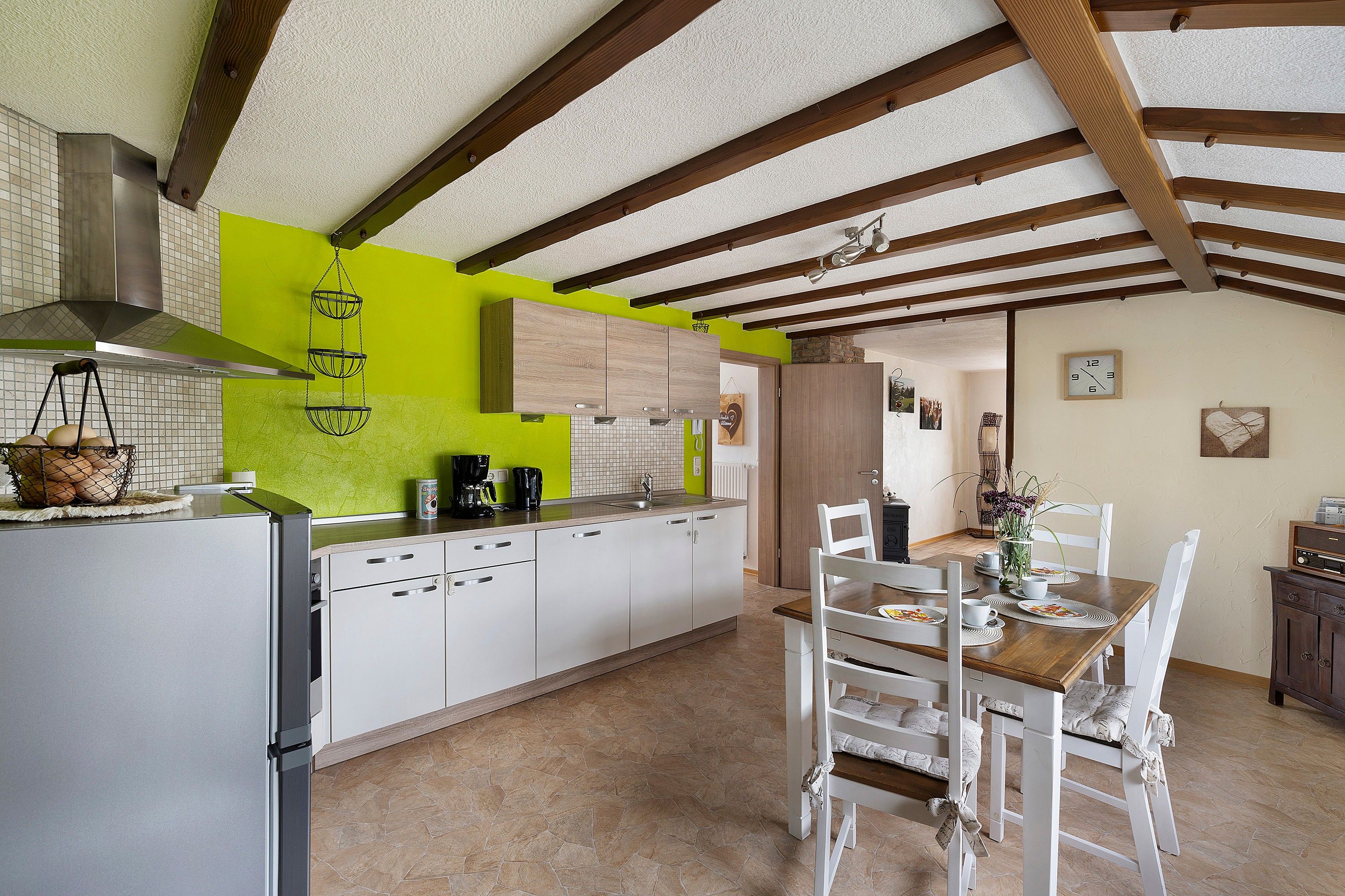 Une cuisine avec des meubles blancs, un mur d’accent vert, un luminaire suspendu et un coin repas avec des chaises blanches et une table en bois.