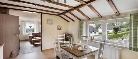 Un coin repas avec une table en bois dressée pour quatre personnes, des chaises blanches et une vue sur l’extérieur par une fenêtre ornée de rideaux.