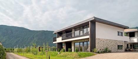 A modern two-story house with a balcony, a gravel driveway, and a mountainous backdrop.