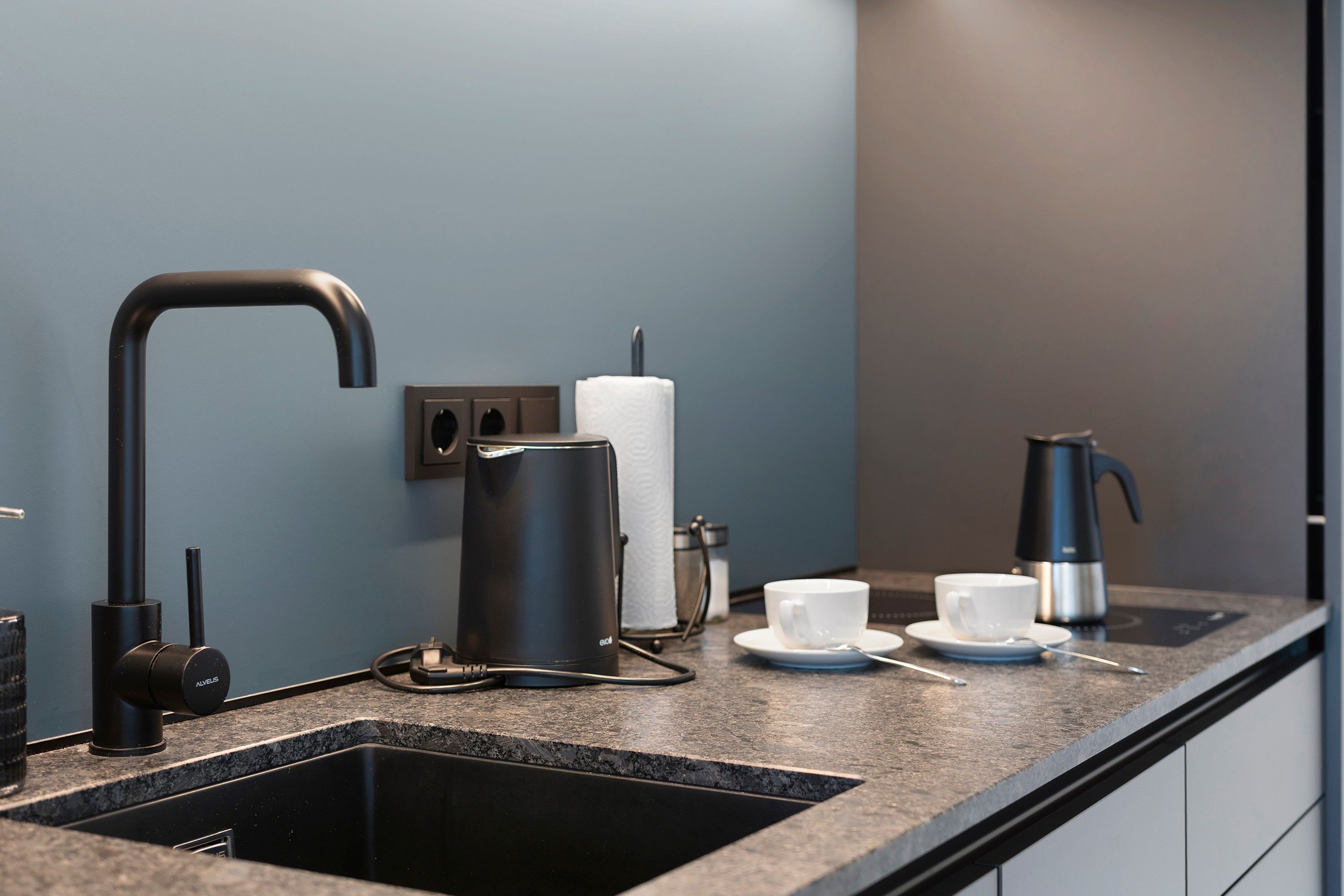 Een moderne keuken met een zwarte spoelbak, een koffiezetapparaat, een waterkoker en een beker op het aanrecht.