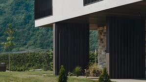 A modern two-story building with a balcony, a stone base, and a view of a forested hill.