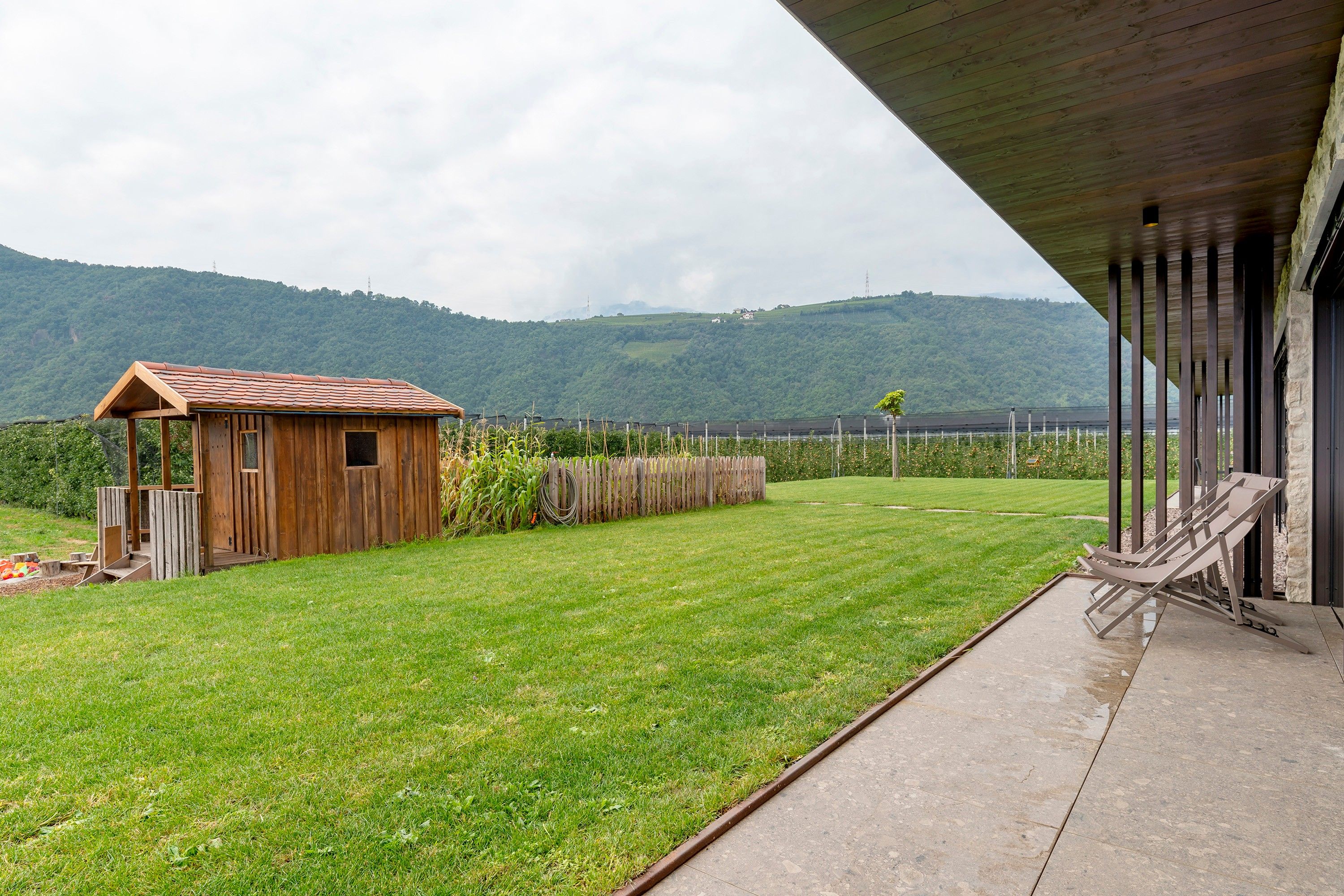 A wooden cabin with a sloped roof, a grassy yard, a paved patio, and a view of a mountain range.