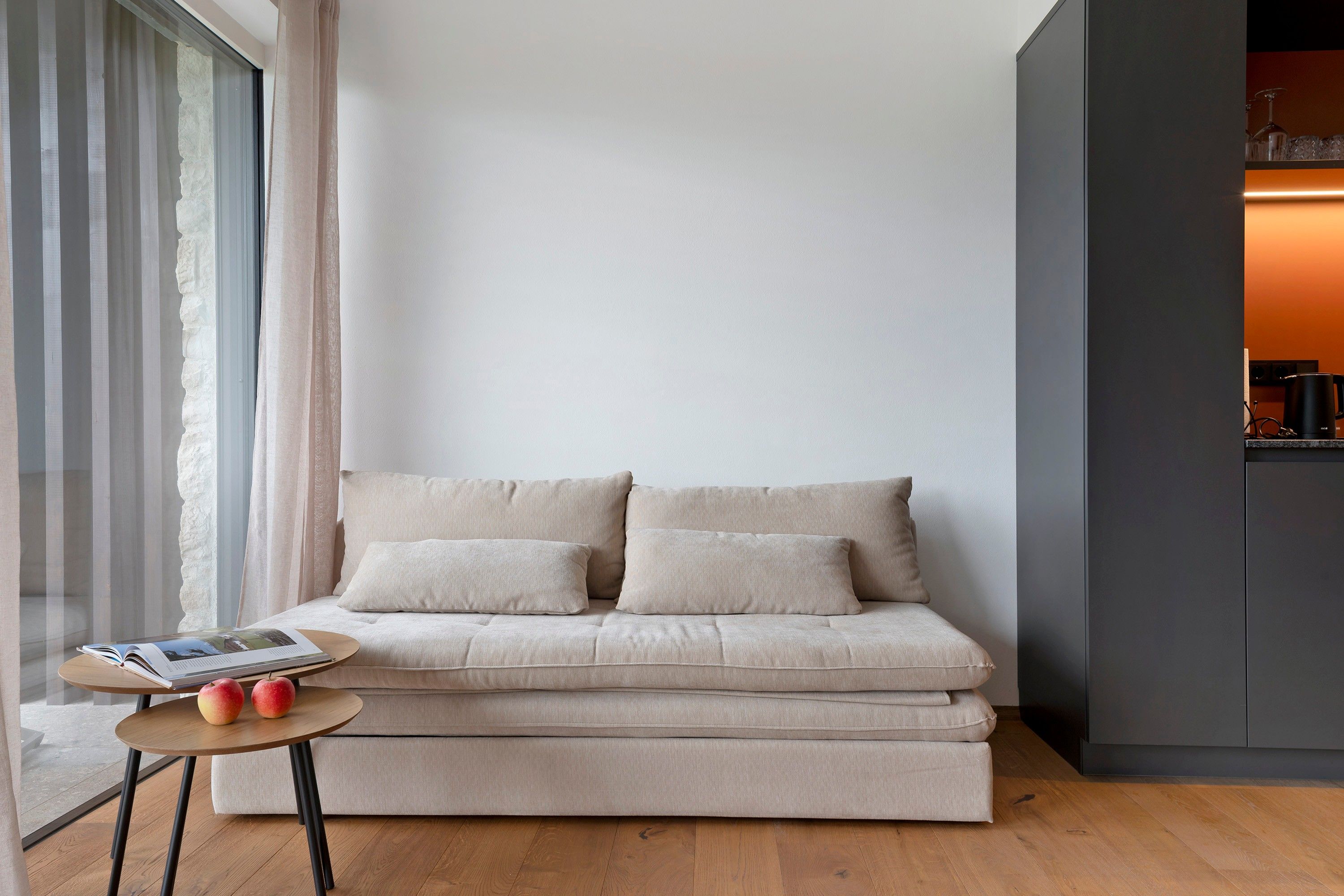 A modern living room with a beige sofa, a small round table with apples, and a view of a kitchen area.