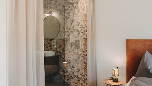 A bedroom with a bed, a nightstand with a lamp, a wooden floor, and a bathroom visible through a curtain.