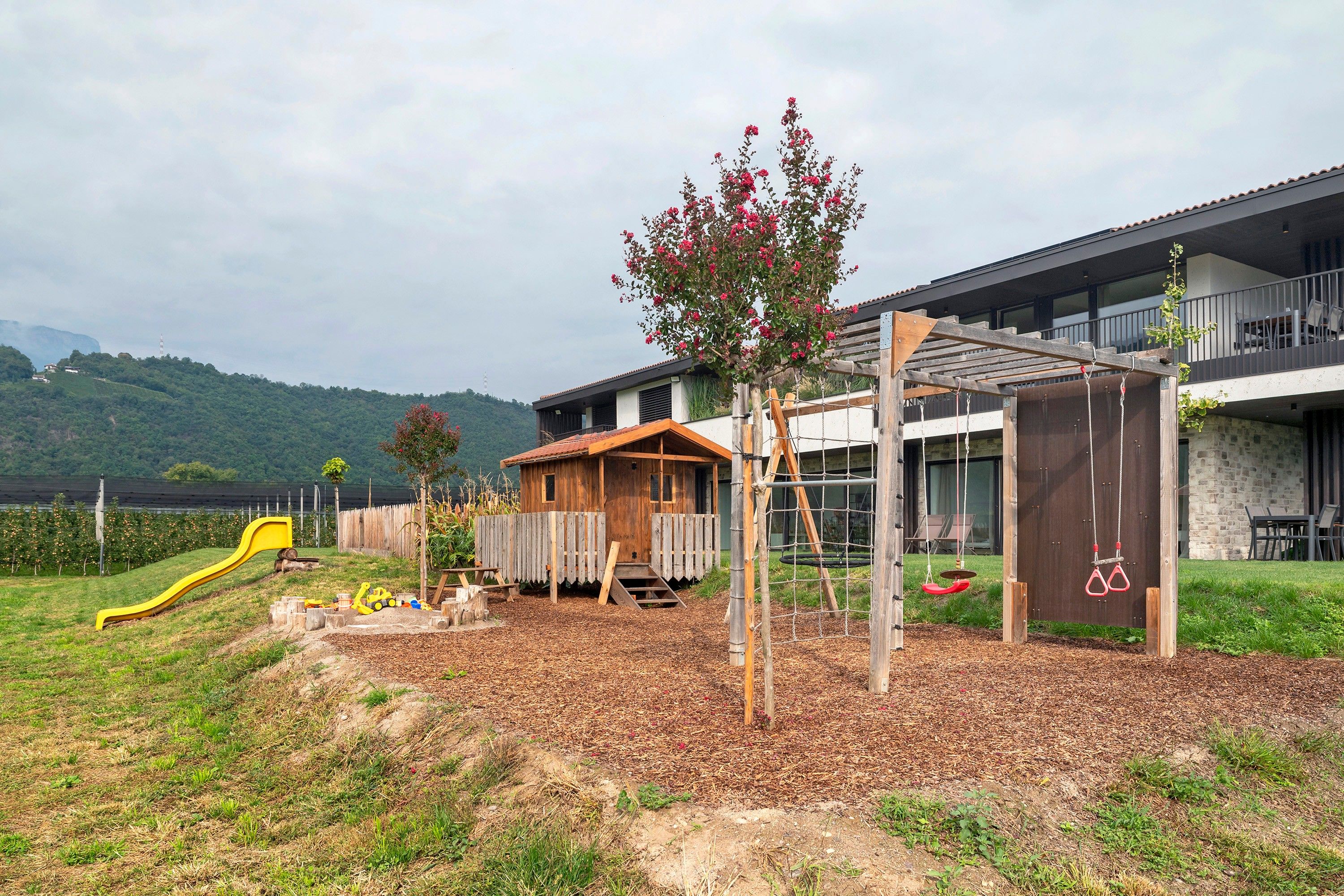 Un terrain de jeux avec un toboggan et des balançoires, une cabane en bois et un bâtiment doté d’un balcon.