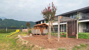Un terrain de jeux avec un toboggan et des balançoires, une cabane en bois et un bâtiment doté d’un balcon.