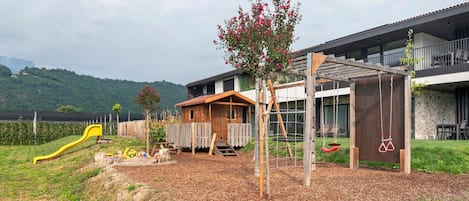 A playground with a slide and swings, a wooden playhouse, and a building with a balcony.