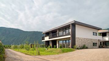 Une maison moderne à deux étages, dotée d’un balcon, d’une allée en gravier et surplombant un paysage montagneux.