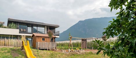 Una casa moderna amb una finestra gran, una caseta de joc de fusta i un tobogan groc al pati.