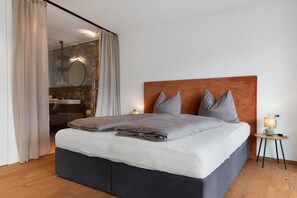Une chambre à coucher moderne avec un grand lit, une table de chevet avec une lampe et une salle de bain visible à travers une porte en verre.