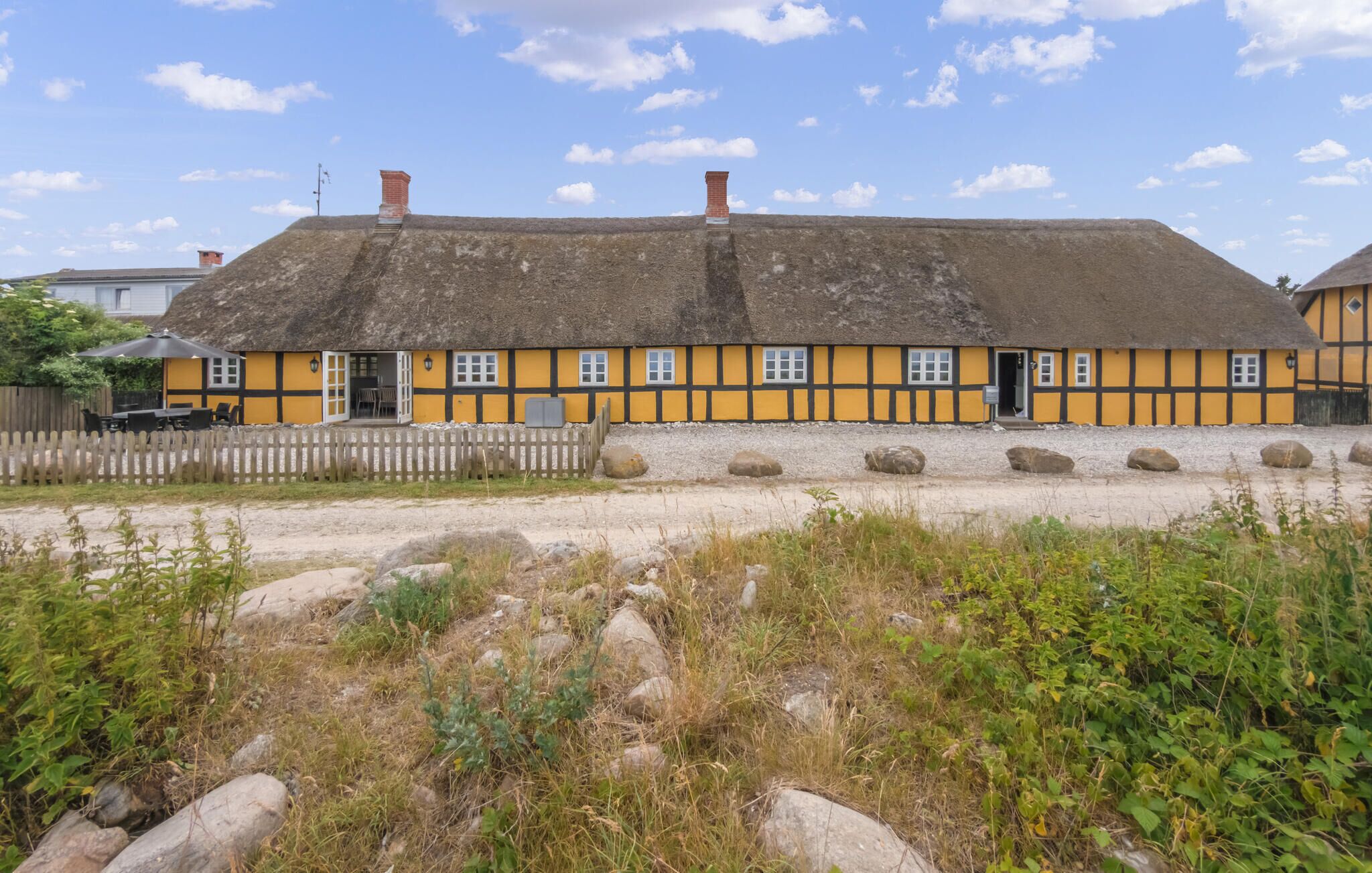 A traditional yellow and black half-timbered house with a thatched roof, a wooden fence, and a gravel path in front.