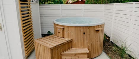 A wooden hot tub with steps leading up to it, surrounded by a white fence and greenery.