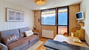 Ein gemütliches Zimmer mit Sofa, Tisch, Fernseher und Fenster mit Bergblick.