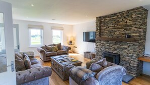 A living room with a stone fireplace, a television mounted on the wall, and a large window. - 5 The Cloisters - Waterville (Waterville)