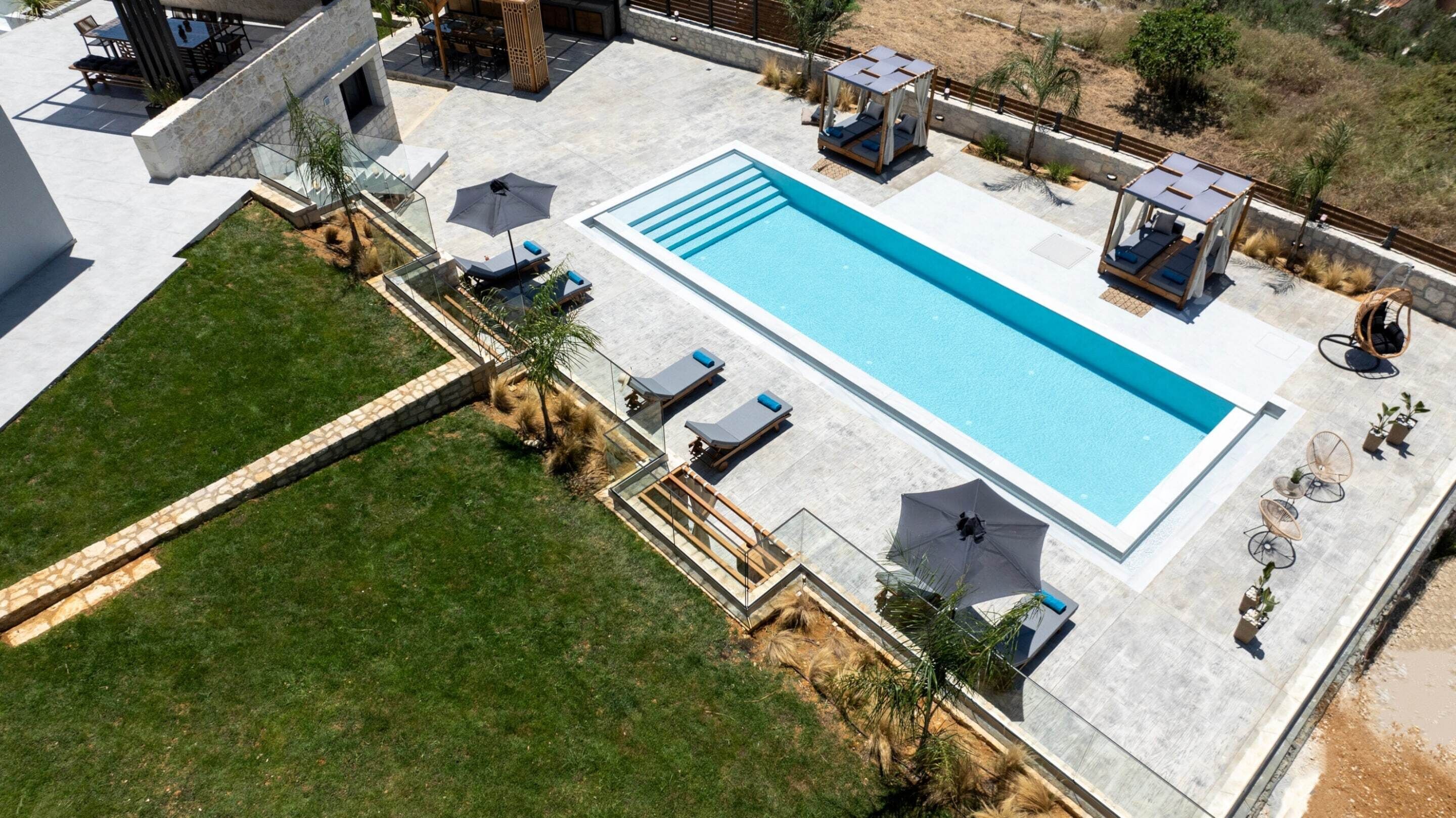 Piscina rectangular con sillones y sombrillas, rodeada por un suelo de piedra y una zona de césped.