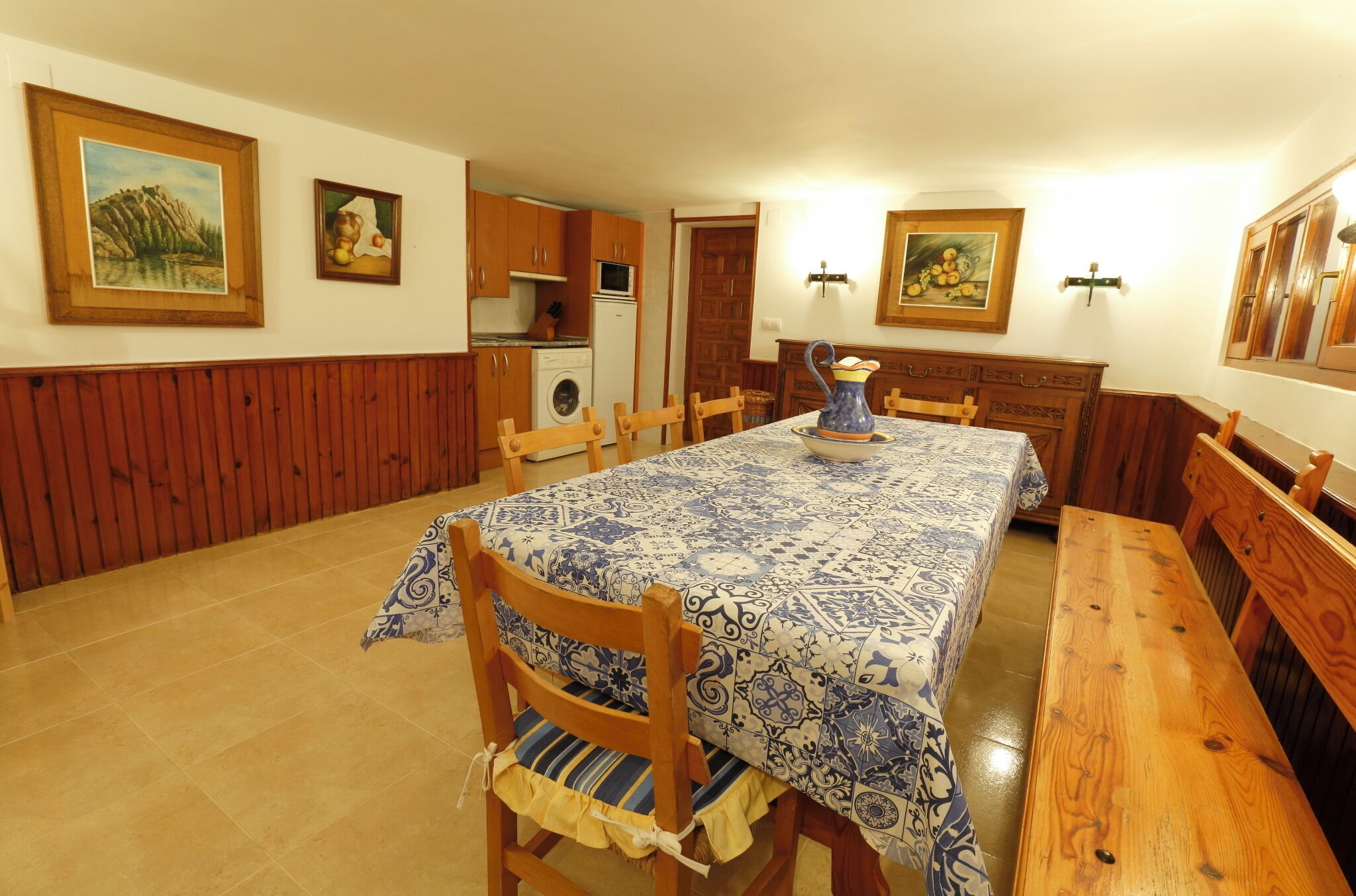 A dining area with a table set for four, wooden chairs, a bench, and a kitchen in the background.