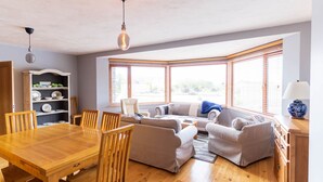 A living room with a wooden dining table, beige sofas, and a bay window with blinds. - Lig do Scíth - Cahersiveen (Cahersiveen)