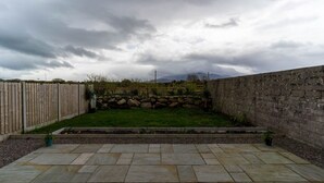 Ein Garten mit einer Steinterrasse, einem Holzzaun, einer Steinmauer und einer Rasenfläche mit Blick auf das Gebirge.