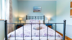 Een bed met een metalen frame, een bloemenbedekker, twee bedlampjes, een aan de muur hangend schilderij en een raam met gordijnen.