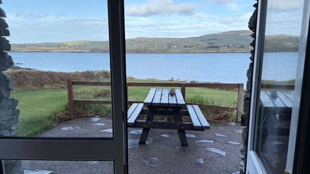 A stone-paved patio with a wooden picnic table and benches, overlooking a body of water and hills. - Skellig Cottage - Portmagee (Portmagee)
