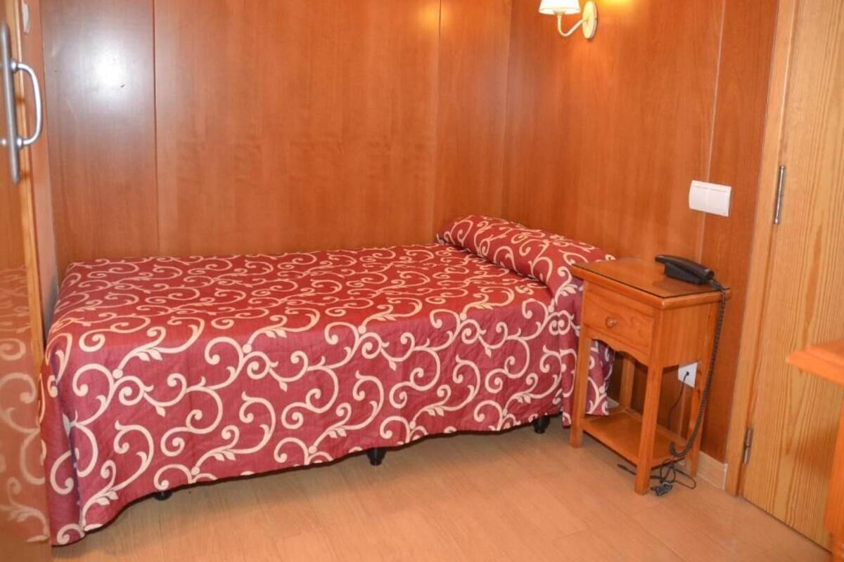 A room with a bed, a bedside table with a phone, and a wall-mounted lamp.