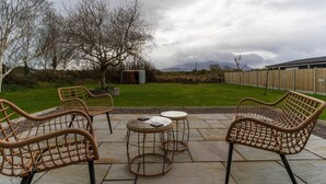 Une terrasse avec des chaises et des tables en osier, un petit abri de jardin et une pelouse offrant une vue sur les montagnes.